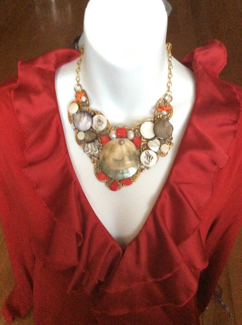 May include: A red ruffled blouse with a large, gold, and white seashell necklace. The necklace features a variety of shells, beads, and buttons.