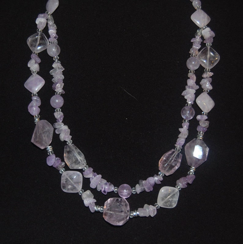 May include: A double-strand necklace made with light purple amethyst and clear quartz beads. The necklace features a variety of shapes and sizes of beads, including round, oval, and rectangular.