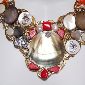 May include: A gold necklace with a large iridescent shell pendant, surrounded by smaller shells, red coral beads, and silver Om symbols.