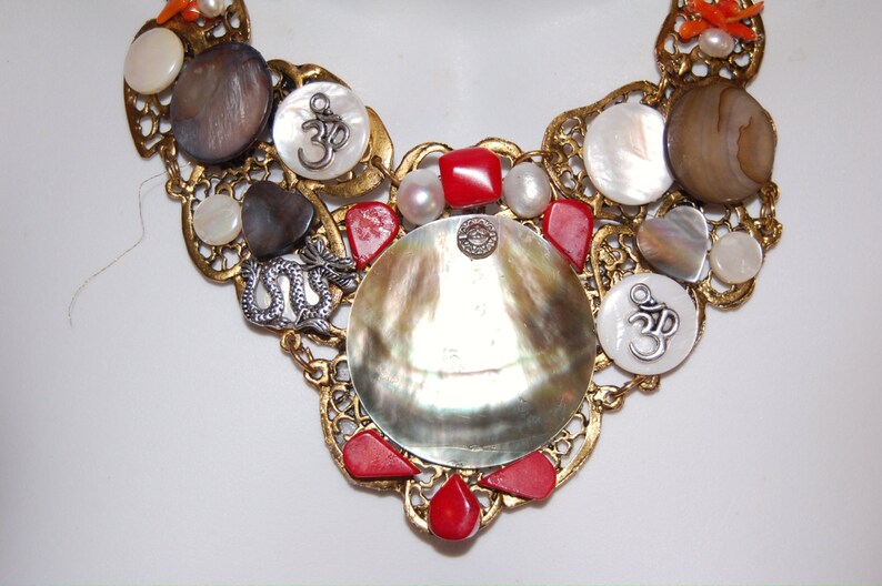 May include: A gold necklace with a large iridescent shell pendant, surrounded by smaller shells, red coral beads, and silver Om symbols.