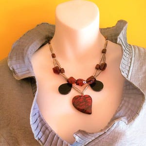 May include: A gray shirt with a layered necklace featuring red and black beads, a heart pendant, and a silver chain.