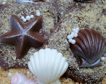 May include: Close-up of chocolate treats decorated with edible starfish, seashells, and white pearl sprinkles. The treats sit on a surface covered in crushed graham crackers, resembling sand. The chocolate is dark brown and the seashells are white.