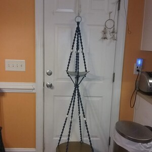May include: A macrame plant hanger with two round wooden shelves. The hanger is made of dark blue cord and has white beads. The hanger is hanging from a door.