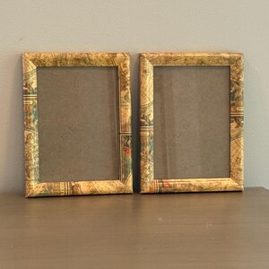 May include: Two rectangular picture frames featuring a vintage map print with muted tones of beige, brown, green, and blue. The frames have a smooth finish and contain clear glass panels, each surrounding a plain, neutral-colored backing board.