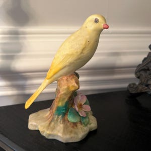 May include: A yellow porcelain bird figurine perched on a branch with pink flowers. The bird has a red beak and black eyes.