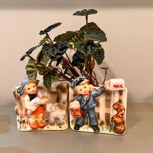 May include: A pair of vintage ceramic planter figurines depicting a girl holding a lamb and a boy delivering mail. The planters are shaped like a fence with a mailbox and a dog on each side. The planters are decorated with green leaves and flowers.