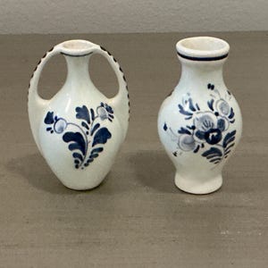 May include: Two small white ceramic vases featuring hand-painted blue floral patterns. The left vase has two arched handles, while the right vase has a classic bulbous shape with a flared rim. Both pieces display intricate blue botanical motifs.