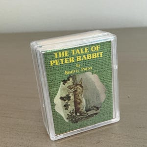 May include: A miniature book titled The Tale of Peter Rabbit by Beatrix Potter, encased in a clear plastic protective sleeve. The cover features a green textured background with yellow text and an illustration of a brown rabbit standing upright.