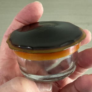 May include: A small, clear glass jar featuring a round, dark brown lid with a scalloped edge. The lid sits atop a vibrant, translucent orange band that connects to the transparent glass base. The overall design is compact and circular.