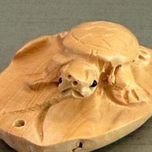 May include: A light tan wooden carving of a turtle resting on a flat, leaf-shaped base. The turtle features a detailed shell, four legs, and two small black eyes. A small circular hole is visible on the left side of the base for hanging purposes.