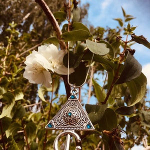 Puede incluir: Un collar de plata con un colgante triangular que presenta intrincados dise&ntilde;os de filigrana y piedras preciosas azules. El collar est&aacute; colgado de una rama con flores blancas.