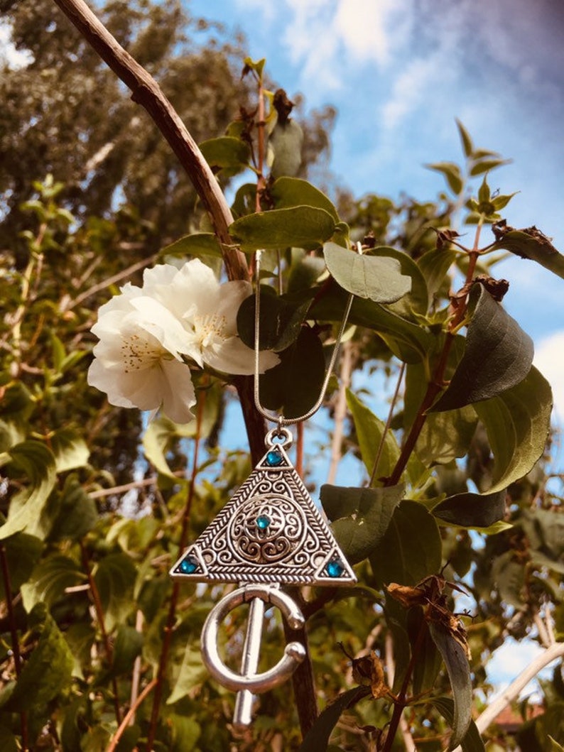 Puede incluir: Un collar de plata con un colgante triangular que presenta intrincados dise&ntilde;os de filigrana y piedras preciosas azules. El collar est&aacute; colgado de una rama con flores blancas.