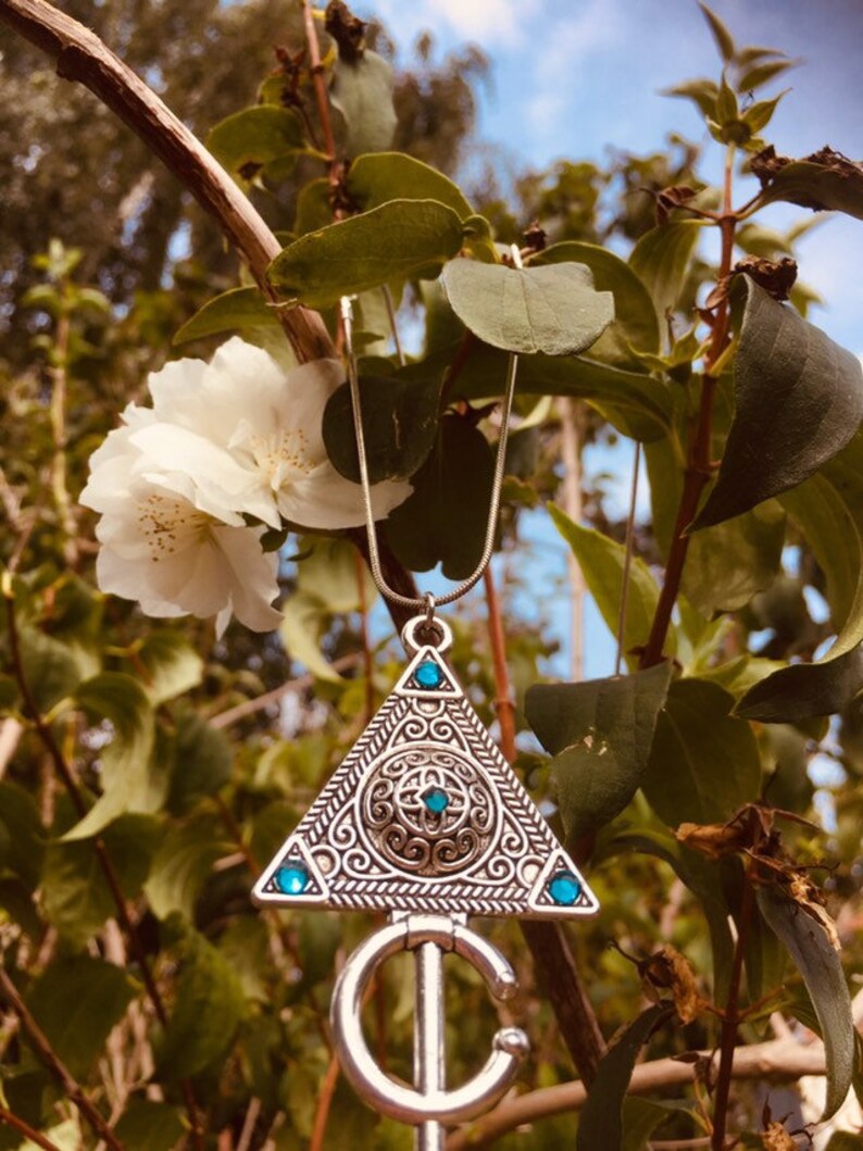 Puede incluir: Un collar de plata con un colgante triangular que presenta un dise&ntilde;o intrincado y piedras preciosas azules. El colgante tiene forma de llave con una cabeza circular y un tallo largo y delgado.