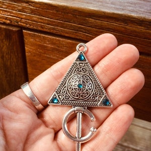 Puede incluir: Un colgante de tri&aacute;ngulo plateado con dise&ntilde;os intrincados y piedras preciosas azules. El colgante tiene un anillo unido a la parte inferior.