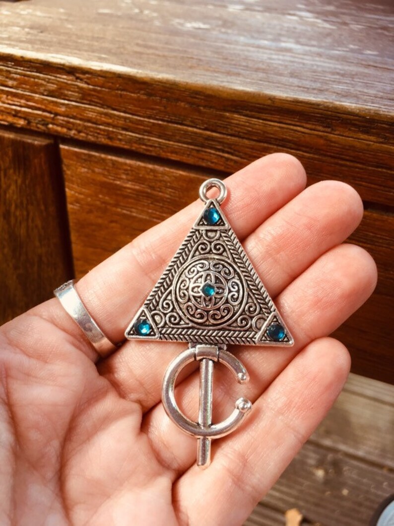 Puede incluir: Un colgante de tri&aacute;ngulo plateado con dise&ntilde;os intrincados y piedras preciosas azules. El colgante tiene un anillo unido a la parte inferior.