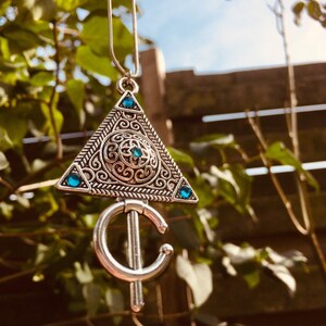 Puede incluir: Un collar de plata con un colgante triangular que presenta intrincados dise&ntilde;os de filigrana y piedras preciosas azules. El colgante est&aacute; unido a una cadena de plata.