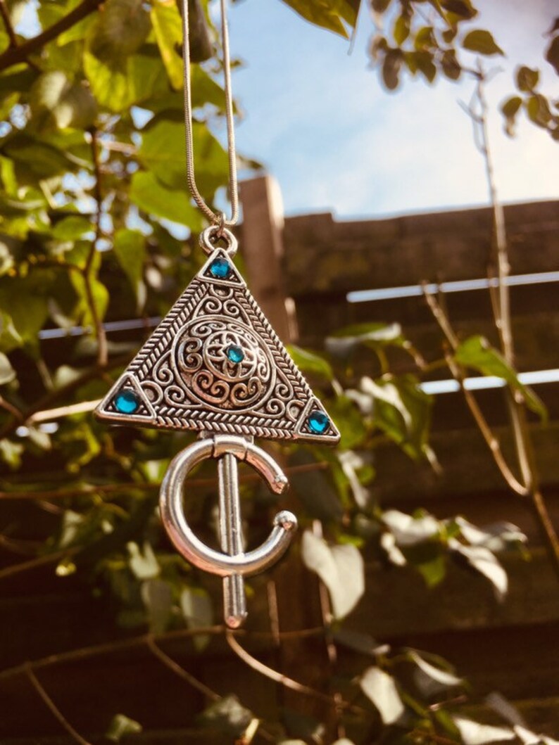 Puede incluir: Un collar de plata con un colgante triangular que presenta intrincados dise&ntilde;os de filigrana y piedras preciosas azules. El colgante est&aacute; unido a una cadena de plata.