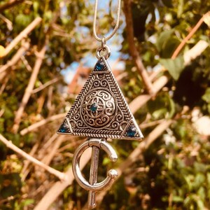 Puede incluir: Un collar de plata con un colgante triangular que presenta intrincados dise&ntilde;os de filigrana y piedras preciosas azules. El colgante est&aacute; unido a una cadena de plata.
