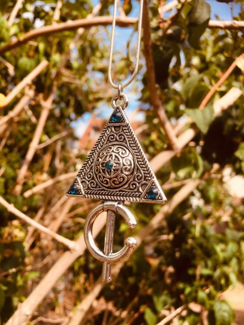 Puede incluir: Un collar de plata con un colgante triangular que presenta intrincados dise&ntilde;os de filigrana y piedras preciosas azules. El colgante est&aacute; unido a una cadena de plata.