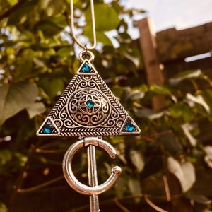 Puede incluir: Un collar de plata con un colgante triangular que presenta intrincados dise&ntilde;os de filigrana y piedras preciosas azules. El colgante est&aacute; unido a una cadena de plata.