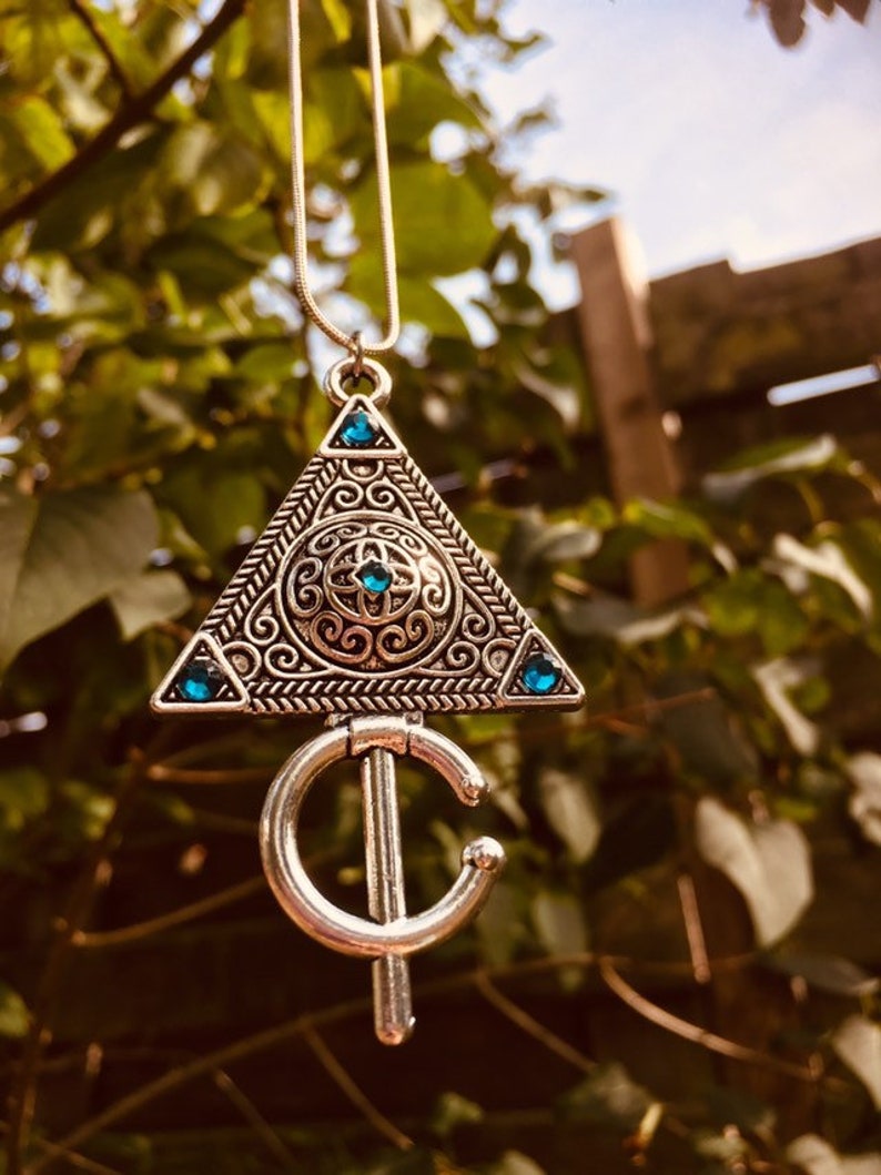 Puede incluir: Un collar de plata con un colgante triangular que presenta intrincados dise&ntilde;os de filigrana y piedras preciosas azules. El colgante est&aacute; unido a una cadena de plata.