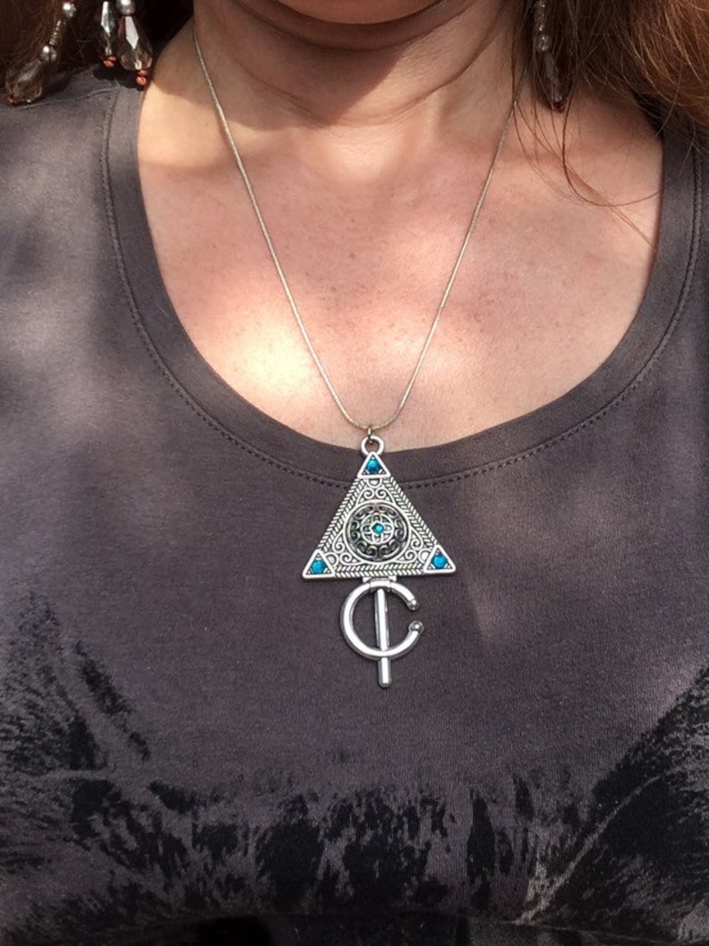 Puede incluir: Un collar de plata con un colgante triangular que presenta un dise&ntilde;o detallado con piedras preciosas turquesa. El colgante est&aacute; unido a una cadena de plata.