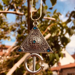 Puede incluir: Un collar con un colgante de plata en forma de tri&aacute;ngulo adornado con un dise&ntilde;o intrincado y piedras preciosas azules. El colgante est&aacute; unido a una forma de media luna de plata.