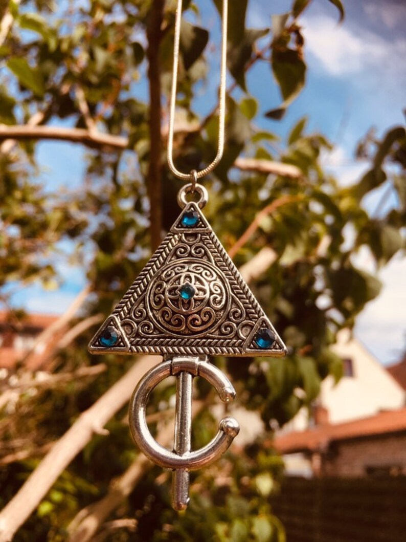 Puede incluir: Un collar con un colgante de plata en forma de tri&aacute;ngulo adornado con un dise&ntilde;o intrincado y piedras preciosas azules. El colgante est&aacute; unido a una forma de media luna de plata.