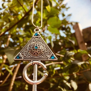 Puede incluir: Un collar de plata con un colgante triangular que presenta dise&ntilde;os intrincados y piedras preciosas azules. El colgante est&aacute; unido a una cadena de plata con un colgante en forma de media luna.