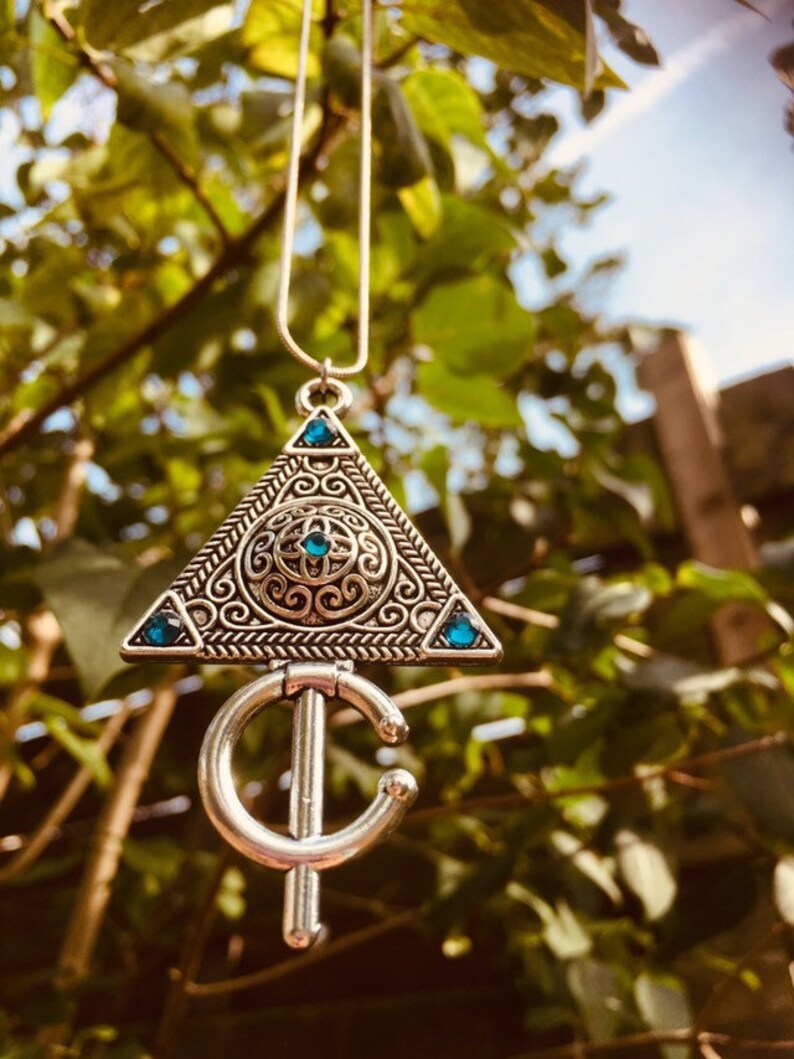 Puede incluir: Un collar de plata con un colgante triangular que presenta dise&ntilde;os intrincados y piedras preciosas azules. El colgante est&aacute; unido a una cadena de plata con un colgante en forma de media luna.