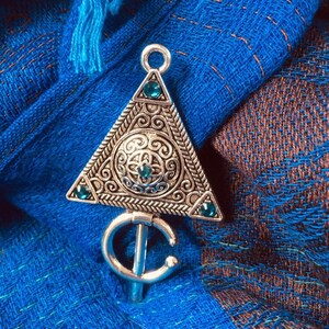 Puede incluir: Un colgante de plata con forma triangular y detalles intrincados. El colgante presenta un dise&ntilde;o circular con una piedra preciosa verde en el centro y piedras preciosas verdes m&aacute;s peque&ntilde;as alrededor de los bordes. El colgante est&aacute; unido a una cadena de plata con un gancho en forma de C.