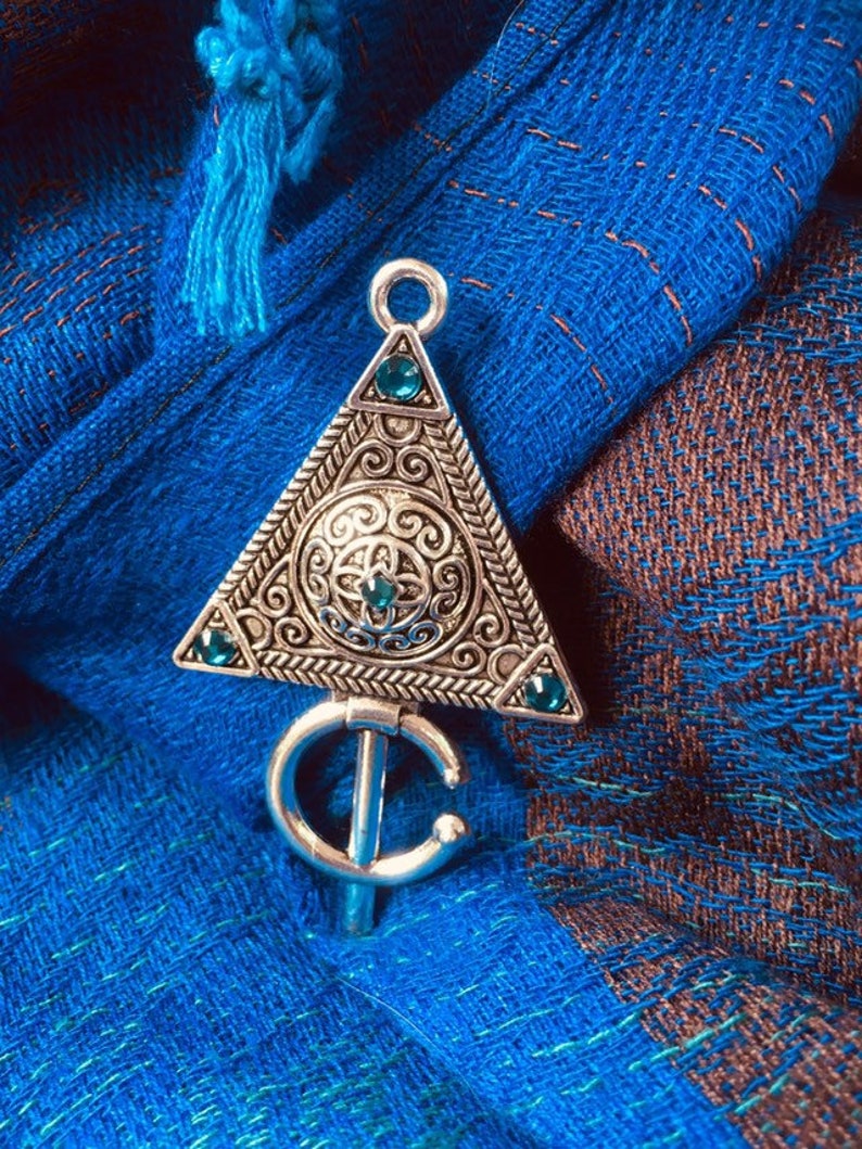 Puede incluir: Un colgante de plata con forma triangular y detalles intrincados. El colgante presenta un dise&ntilde;o circular con una piedra preciosa verde en el centro y piedras preciosas verdes m&aacute;s peque&ntilde;as alrededor de los bordes. El colgante est&aacute; unido a una cadena de plata con un gancho en forma de C.