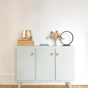 Peut inclure: Un buffet bleu clair avec trois portes et des boutons dorés. Le buffet a un design simple avec des lignes droites et un dessus légèrement arrondi. Le buffet est posé sur un sol en bois. Il y a deux paires de chaussures dorées sur le dessus du buffet.