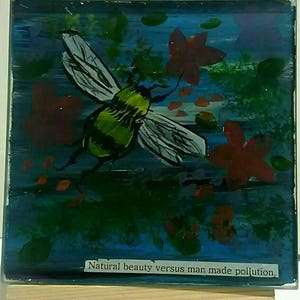 May include: A small painting on a wooden easel depicts a bee with yellow and green stripes flying over red flowers against a blue background. The painting has the text "Natural beauty versus man made pollution."