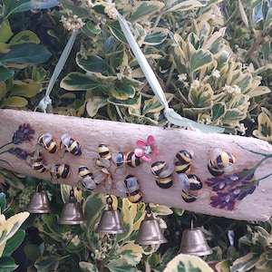 May include: A rectangular piece of light-colored driftwood hanging from a pale green ribbon. The wood is decorated with painted lavender sprigs and several small, three-dimensional bumblebee figures. Five small brass bells hang from the bottom.