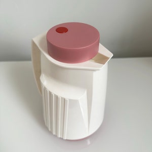 Peut inclure: Une carafe isotherme blanche avec un couvercle rose et un bouton rouge sur le dessus. La carafe a un design côtelé et un bec verseur sur le côté.