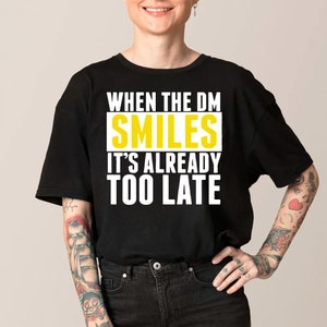 May include: Black t-shirt with the text "WHEN THE DM SMILES IT'S ALREADY TOO LATE" in white and yellow. The shirt is a casual style, suitable for everyday wear. The text is centered on the front of the shirt.
