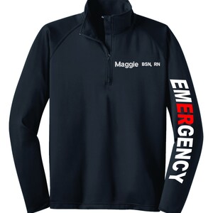 May include: A navy blue long-sleeve performance shirt with a white zipper and a white embroidered name tag that reads "Maggie BSN, RN". The right sleeve has the word "EMERGENCY" printed vertically in white with the letter "E" in red.
