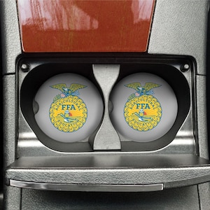 May include: Two gray round car coasters with a blue and yellow FFA logo. The logo features a golden eagle with a banner that reads "FFA" and the words "Agricultural Education" around the outside.