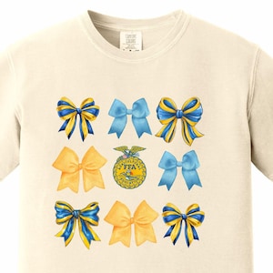 May include: A cream-colored t-shirt with a blue and yellow bow design. The center of the shirt features a blue and yellow FFA emblem.