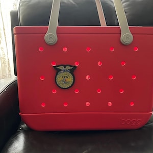May include: A bright red tote bag with a light gray handle and a decorative FFA emblem. The bag has multiple circular cutouts and the brand name "bogg" is visible on the bottom right.