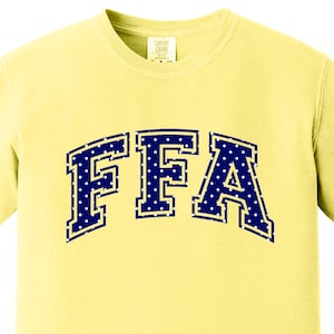 May include: A pale yellow short-sleeve t-shirt featuring the letters FFA in a dark blue collegiate-style font. The letters are filled with a white polka dot pattern and outlined in white, positioned in an arched layout across the chest.