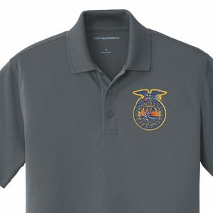May include: A charcoal gray polo shirt with a collared neckline and short sleeves. The shirt features a patch with a gold border and a design with an eagle and the letters FFA. The shirt is made of a soft, breathable fabric.