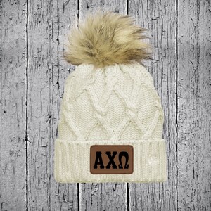 May include: White cable knit beanie with a brown faux fur pom pom and a brown leather patch with the letters "AXO" in black.