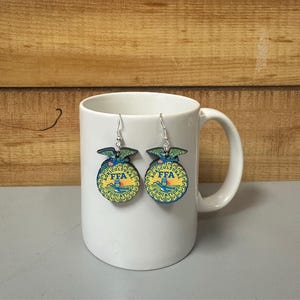 May include: A pair of dangle earrings featuring the FFA emblem. The earrings have a circular design with a yellow background, blue and green accents, and the letters "FFA". The earrings are displayed on a white mug.