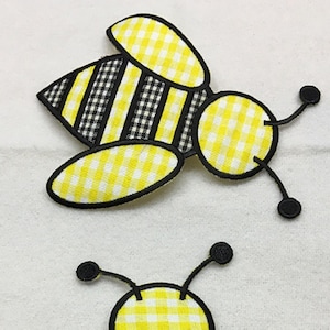 Op de afbeelding: Twee gele en zwarte bijenapplicaties met een ruitjespatroon. De bijen zijn zwart omrand en hebben zwart-wit gestreepte lichamen.