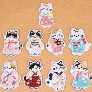 May include: A collection of embroidered cat patches in various colors, each wearing a traditional Japanese kimono. The patches feature cats with black, white, and orange fur, and the kimonos are in shades of pink, blue, and red.