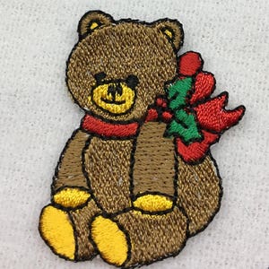 May include: A brown teddy bear wearing a red scarf and a green and red holly sprig. The bear is sitting with its legs crossed and has a yellow nose and eyes.
