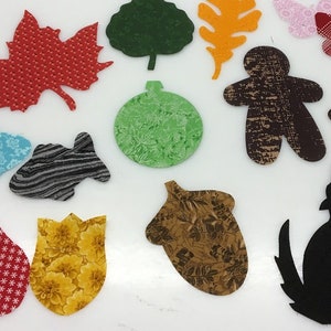 May include: A collection of fabric cutouts in various shapes and patterns. Includes a red maple leaf, green tree, orange leaf, gingerbread people, butterflies, fish, acorn, and a howling wolf. The cutouts have diverse textures and colors, suitable for crafts.