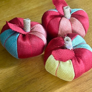 May include: Three fabric pumpkins featuring vertical panels in shades of red, teal, and light green. Each pumpkin is topped with a short, wrapped twine stem and a single fabric leaf. The pumpkins have a rounded, segmented shape with visible stitching.